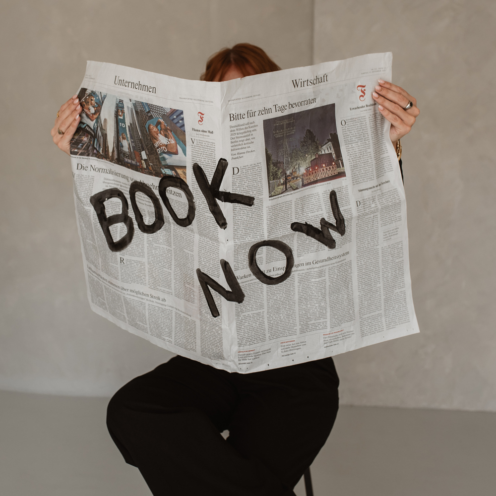 Person sitzt auf Hocker, hält eine Zeitung in der Hand. Auf der Zeitung steht: Book now!